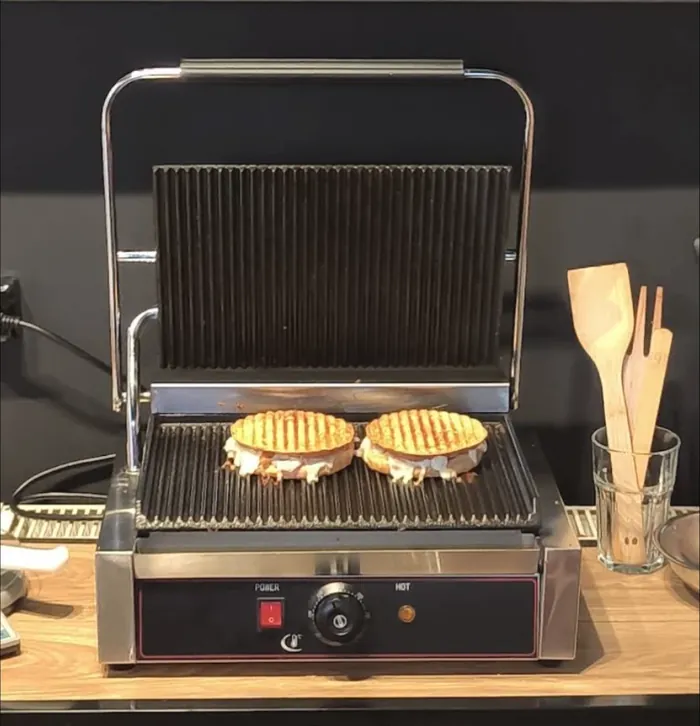 Otwarty grill kontaktowy na drewnianym blacie, na ruszcie dwa tosty z wypływającym serem. Obok szklanka z drewnianymi łopatkami.
