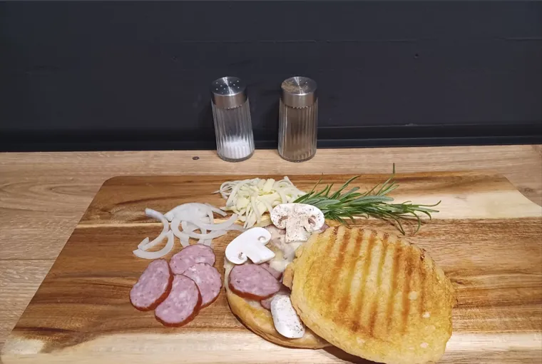 Zapieczony tost na desce, obok plastry kiełbasy, krążki cebuli, plasterki pieczarek, starty ser i gałązka rozmarynu.