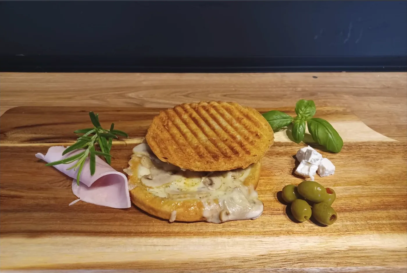 Zapiekany tost z chrupiącą, złocistą skórką w paski z grilla, przekrojony — w środku roztopiony ser i plastry pieczarek. Leży na drewnianej desce, obok gałązka rozmarynu, listki bazylii, zwinięty plaster szynki, kostki fety i zielone oliwki.