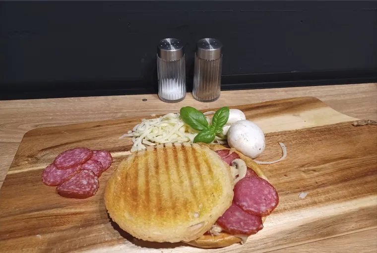 Zapieczony tost, obok na desce wachlarz plastrów salami, starty ser, świeża pieczarka i listki bazylii.