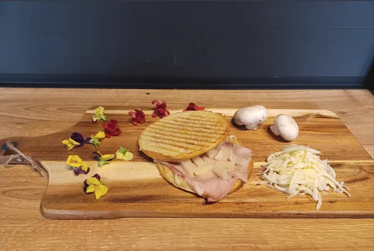 Otwarty tost z plastrami szynki i kostkami ananasa, na drewnianej desce; obok kopczyk startego sera, dwie pieczarki i rozsypane jadalne kwiaty.
