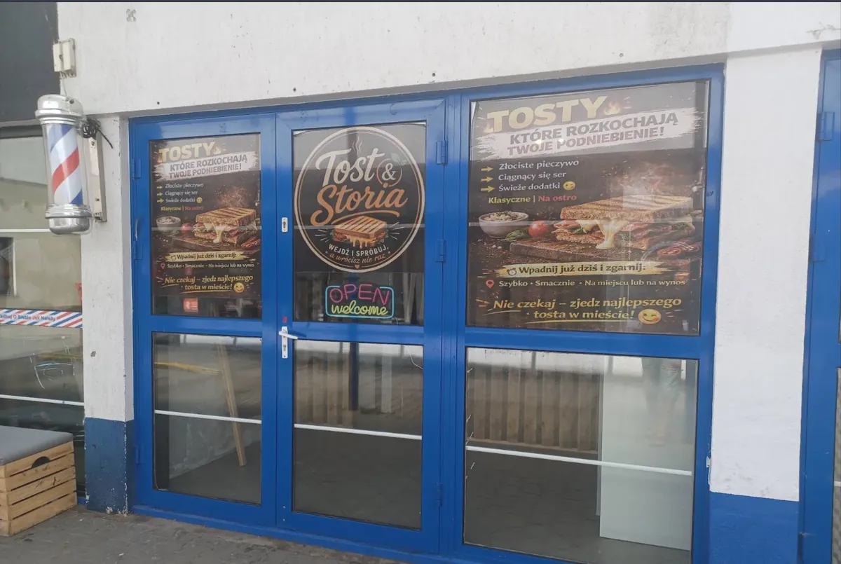 Niebieskie, szklane drzwi w białej ścianie, oklejone dużymi plakatami z napisem TOSTY i okrągłym logo Tost&Storia. Po lewej stronie drzwi, na ścianie, pasiasty słupek fryzjerski. Przed wejściem drewniana ławka.